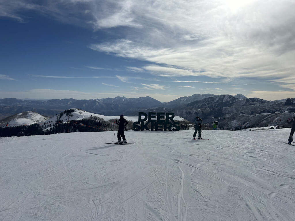 Skiers beside the large Deer Skiers sign on a Deer Valley ridge with the Wasatch Mountains behind