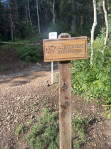 Wooden trailhead sign reading "All Trails Lead to Happiness" beside a gravel path and aspen trees