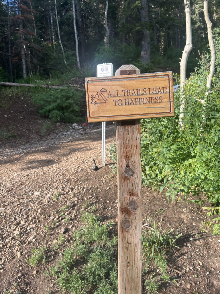 Wooden trailhead sign reading "All Trails Lead to Happiness" beside a gravel path and aspen trees