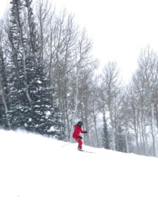 Skier in a red suit carving through fresh snow beside aspen and evergreen trees at Deer Valley