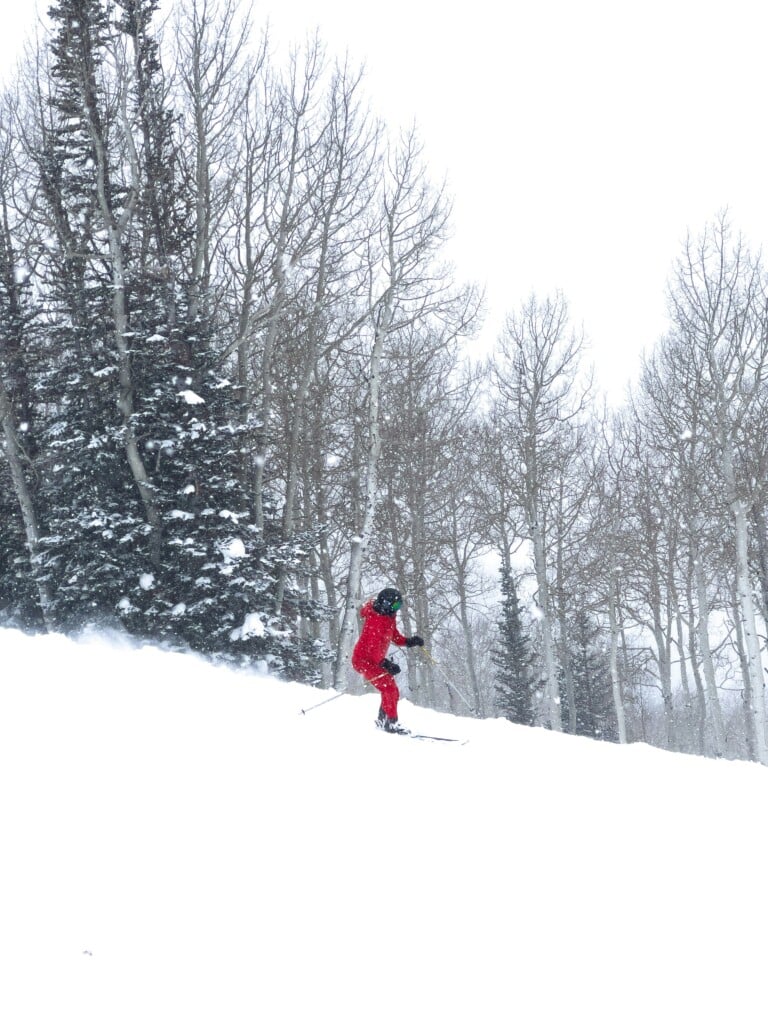 Skier in a red suit carving through fresh snow beside aspen and evergreen trees at Deer Valley